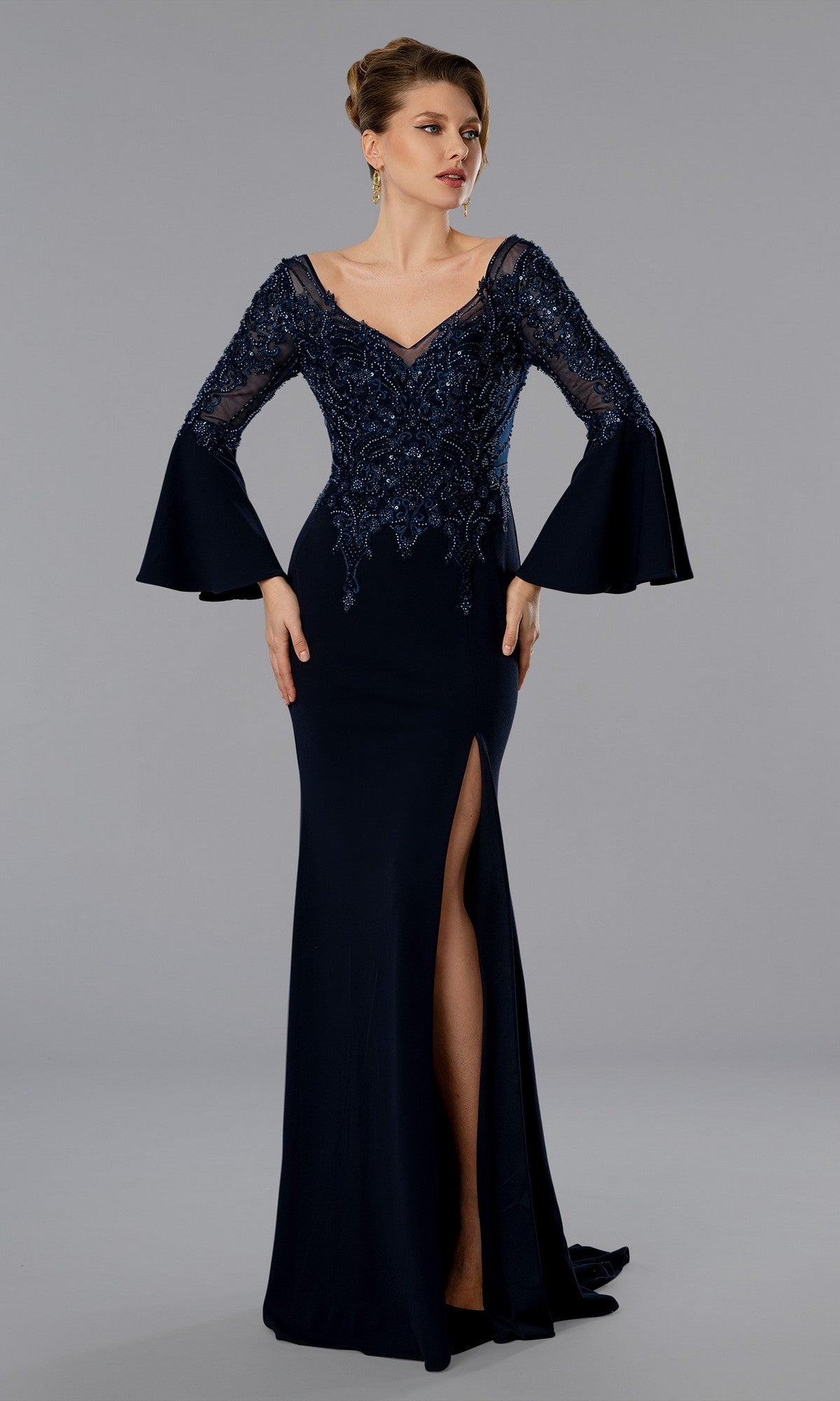 A woman stands against a gray background wearing the Stella Couture 25366 long formal dress in navy, featuring lace detailing, bell sleeves, a V-neckline, beaded bodice, and a thigh-high slit.